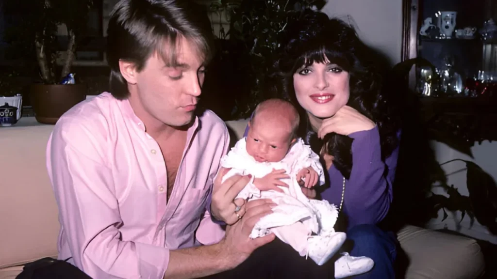 A man in a pink shirt and a dark-haired woman in purple sitting together on a sofa, holding a newborn baby.
