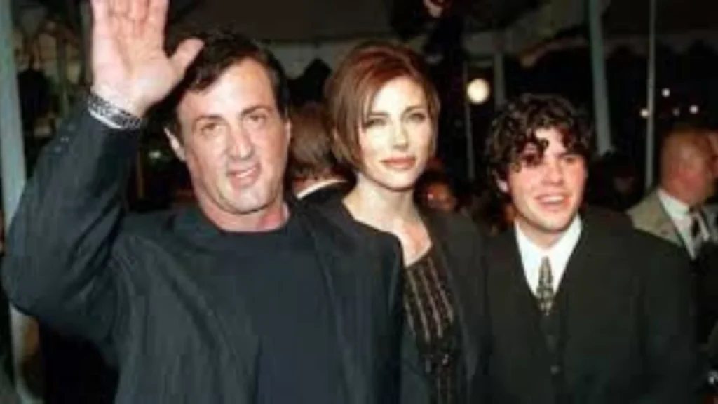 Three people dressed in formal attire at a red carpet event, with one man raising his hand while posing for the camera alongside a woman and a younger man.
